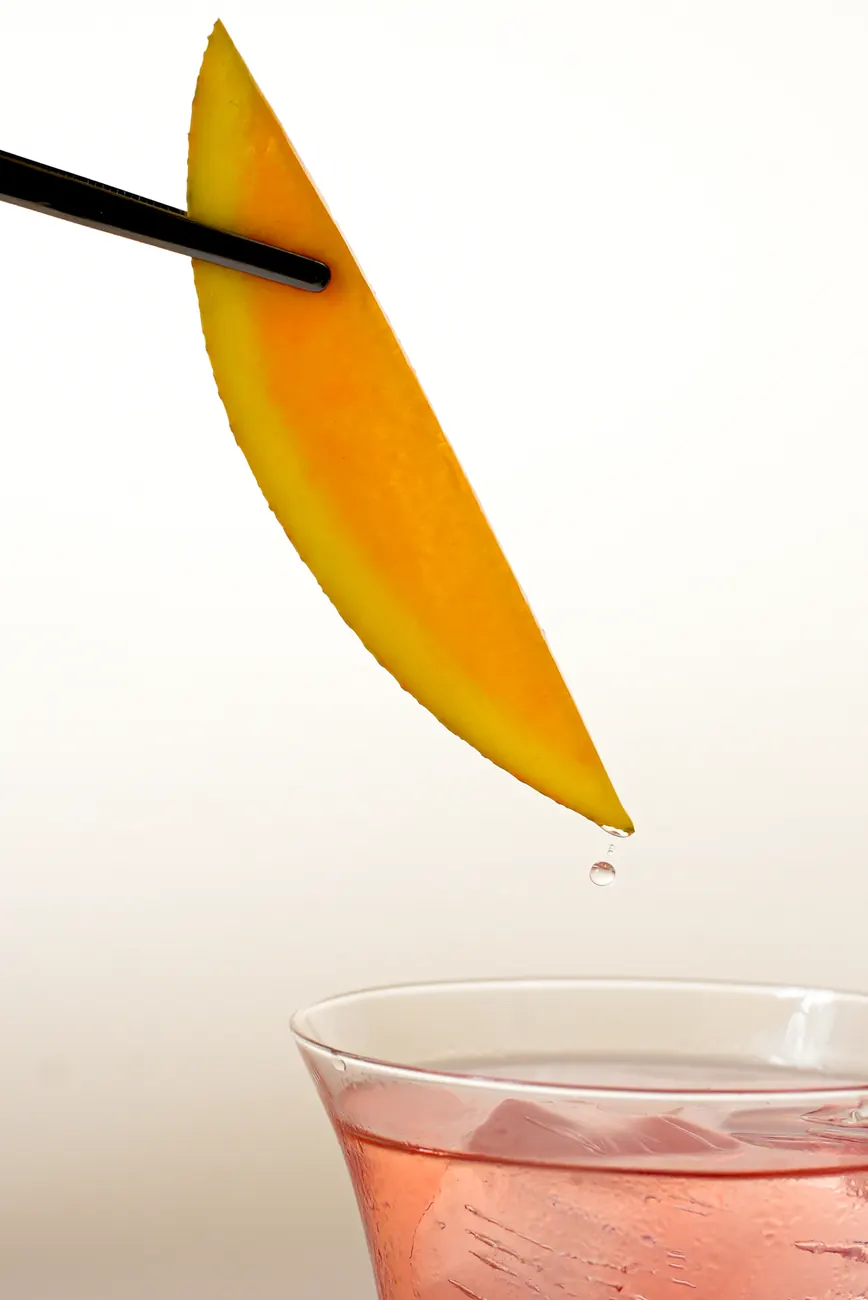 A mango slice on a black skewer above a pink drink