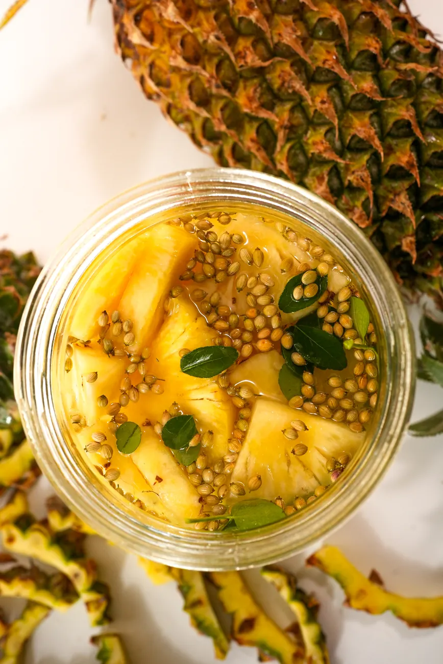 Pineapple pieces in a jar with coriander seeds and leaves, beside a whole pineapple